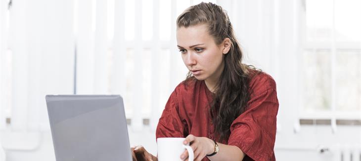 Eine junge Frau sitzt am Laptop und blickt konzentriert auf den Bildschirm. In einer Hand hält sie eine Tasse.