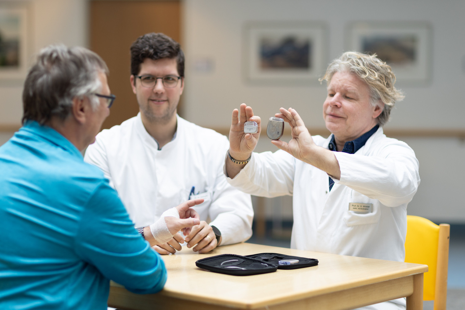 Prof. Dirk Winkler (re.), Leitender Oberarzt und stellvertretender Klinikdirektor, und Dr. Martin Vychopen (Mi.) zeigen ihrem Patienten Uwe Dienst noch einmal das neue Generator-Modell, rechts in der Hand, und einen älteren Vorgänger im Größenvergleich. Das ältere Modell war batteriebetrieben und nicht wiederaufladbar. Das UKL kann als erster in Ostdeutschland den modernsten Generator implantieren.